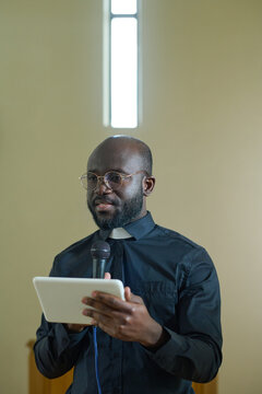 Pastor With Microphone And Tablet Using Electronic Or Online Bible While Pronouncing Sermon In Front Of Parishioners During Church Service