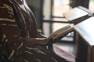 Side view of young black woman in casual dress sitting in church with open Holy Bible while reading verses from Gospel to herself