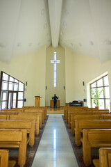 Long aisle between two rows of wooden benches leading towards priest standing in front of altar and looking at cross with his arms raised