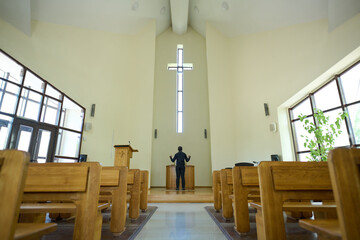 Interior of church with long aisle leading to pastor standing in front of cross and wooden pulpit and keeping his arms in blessing gesture - obrazy, fototapety, plakaty