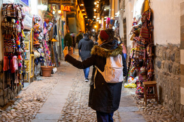 Tourist woman walking through Cusco street at night between shops with cell phone in hand - obrazy, fototapety, plakaty