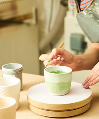 Close-up of girl painting clay mug with glaze. Woman coloring pottery in workshop with a paintbrush. Painter in green apron glazing clay pot. - 516641810