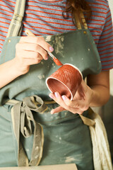 Close-up of girl painting clay mug with glaze. Woman coloring pottery in workshop with a paintbrush. Painter in green apron glazing clay pot.