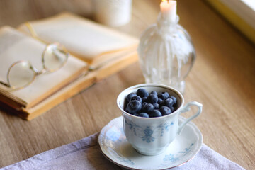 Vintage porcelain cup filled with fresh blueberries, pressed flowers, open book, reading glasses and lit candle on the table. Hygge at home. Selective focus.