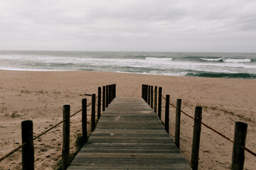 Wooden bridge leads to the ocean waves and sandy clean beach in spring day. Blue sky with clouds and fog. Portugal 