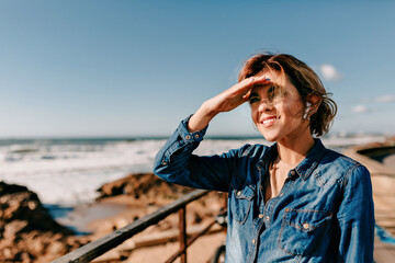 Smiling happy european woman wearing denim shirt standing on ocean shore with on ocean waves background and looking straight while listening music 
