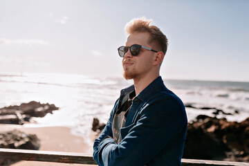 Profile close up portrait of handsome stylish european man in sunglasses wearing dark shirt posing on background of ocean waves in sunlight. Cool guy walking outdoors.