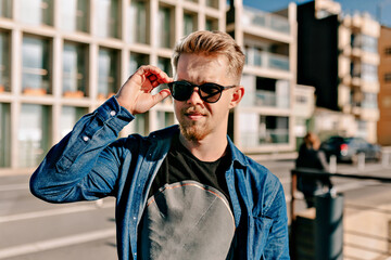 Handsome stylish blond man in sunglasses wearing dark t-shirt and denim shirt posing in sunlight on the shore go ocean in spring day. Man touching glasses and smiling