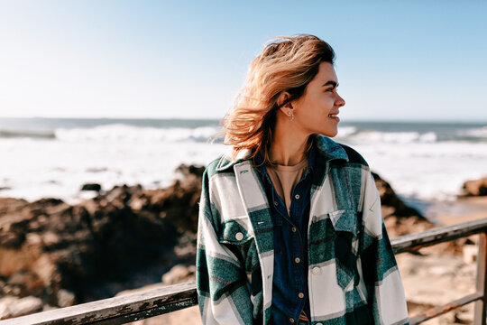 Profile portrait of smiling adorable girl with loose hair looking aside and smiling while standing on background of ocean waves in sunlight. Outdoor photo of european girl in checkered shirt 