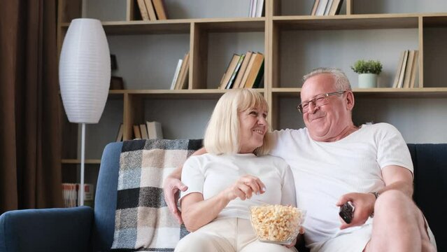 Happy elderly married couple watching tv sitting on sofa, old couple eating popcorn and laughing