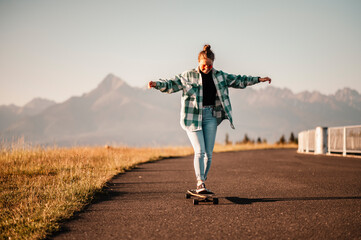 Fototapeta na wymiar Woman rides at straight road on longboard at sunset time. Skater in casual wear training on board during evening sunset with orange light. Girl hold longboard in hands