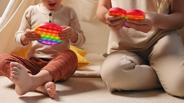 Toddler boy and his mom plays multi-colored toy antistress pop it in children's room near wigwam with soft pillows. Sensory toy rainbow, close-up of the baby's hands having fun with game