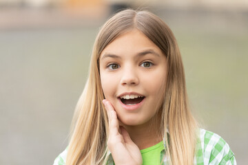 Fototapeta premium Portrait of tween girl with surprised face holding hand on cheek blurry outdoors