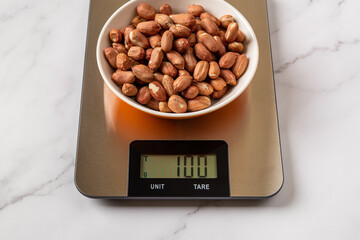 Raw dried peanut in a bowl on a digital kitchen scales. Weigh 100 g whole peeled monkey nut for...