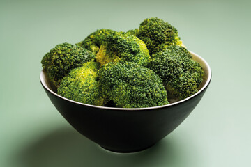 Raw broccoli florets in a black bowl on a turf green colored background. Vegetarian vitamin food closeup. Low calorie antioxidant detox dieting concept. Eat fresh vegetables.