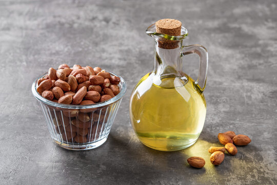 Peanut oil in a glass jug and raw peeled groundnut in a glass bowl over gray background. Arachis hypogaea as edible seeds and oil crop. Monounsaturated cooking oil. Vegetarian snack.