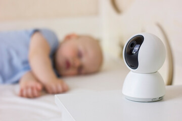 Cute little baby boy sleeping on bed at home with baby monitor camera. - obrazy, fototapety, plakaty