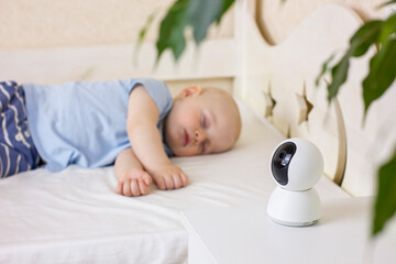 Cute little baby boy sleeping on bed at home with baby monitor camera.