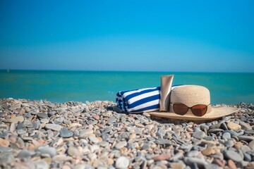 Fototapeta na wymiar A hat, sunglasses, a sun cream and a towel is laying on a stony beach. Blue turquoise Mediterranean sea with waves on the background. High quality photo. 