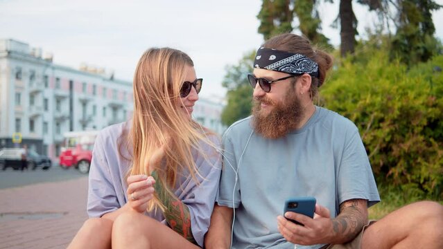 Young cheerful man and women looking a phone screen, put headphones in ears. Smiling diverse friends using smartphone and communicating, listen music, podcast or video chat while sitting on grass park