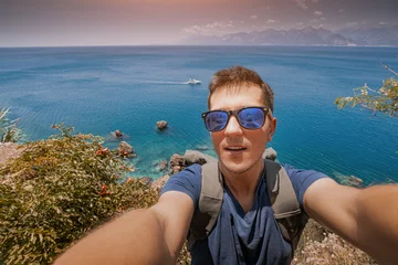 Gordijnen Hiker man with backpack taking selfie photo on a high cliff with splendid view of panoramic seascape and mountains. © EdNurg