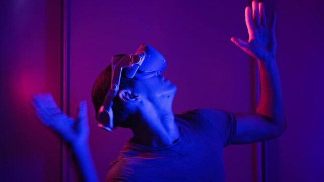 Man in a virtual reality helmet illuminated in red and blue plays a game. Young man in VR headset moves his hands in virtual space. Futuristic technology to play simulation 3D video games Metaverse 4K