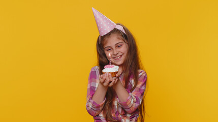 Happy young preteen child girl kid wears festive birthday hat hold cupcake makes wish joyful congratulating blowing burning candle on cake. Little toddler children celebrating anniversary party alone