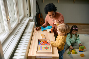 Happy black teacher and small boy playing with puzzle shapes at kindergarten.