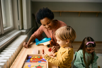 Happy boy playing with colorful puzzle shapes with his kindergarten teacher.