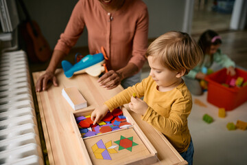 Small boy playing with wooden puzzle shapes with his teacher at kindergarten.