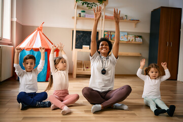Happy black preschool teacher and group of kids stretching during exercise class.