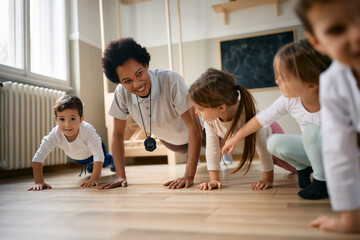 Group of preschool kids and their black female teacher practicing push ups during exercise class.