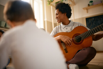 Happy black preschool teacher plays acoustic guitar during music class.