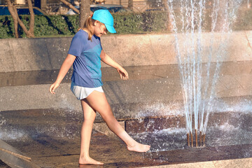 The child is saved from the heat in the fountain. Girl 10-12 years old is cooled by water splashes....