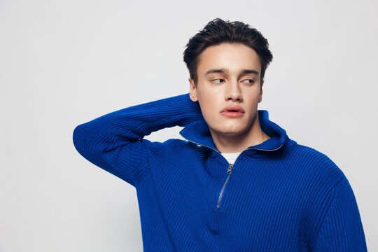 a photo of a beautiful, trendy man with thick black hair standing on a light gray background in a blue zip-up sweater and charmingly looking into empty space.
