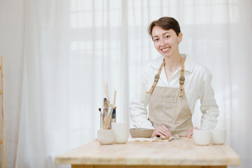 woman ceramist in the workshop makes mugs out of clay. a small business or hobby is the creation of ceramic products. an oriental young woman ceramist makes dishes and vases out of clay. handmade