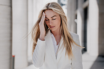 Frustrated blonde caucasian young woman walking eyes closed with upset facial expression touching head, depressed, having financial problems. Beautiful swedish woman having headache outdoors.