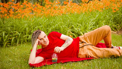 Young man drinking refreshing lemonade, lying on rug in city park. Relaxed male with dreadlocks...