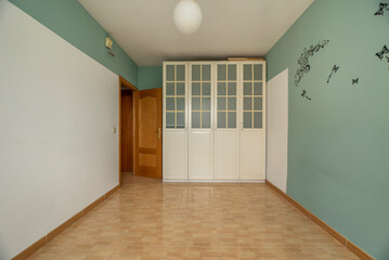 Empty bedroom with wood-like stoneware floors, green and white painted walls and a four-door wood and glass wardrobe