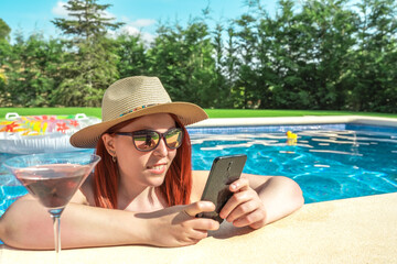 Fototapeta premium young red-haired woman relaxing by the pool, drinking a cocktail and looking at her smartphone. girl on holiday sharing the moments on her social networks. summer concept and free time.