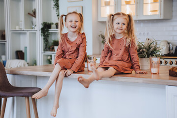 Two Scandinavian cute little sisters with ponytails sitting on kitchen table hugging knees looking at camera. Little swedish girls in pyjamas  on breakfast time at home. Family domestic activities. - obrazy, fototapety, plakaty