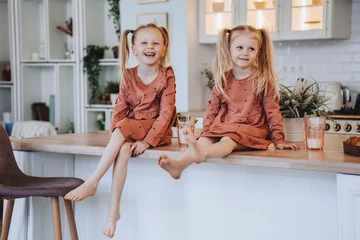 Foto op Aluminium Two Scandinavian cute little sisters with ponytails sitting on kitchen table hugging knees looking at camera. Little swedish girls in pyjamas  on breakfast time at home. Family domestic activities. © Iona