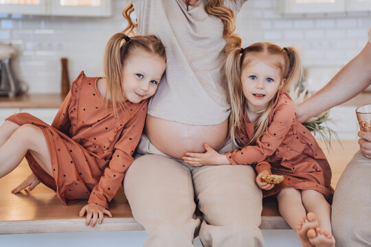 Cropped shoot of two little blonde girls with tails hugging their pregnant mothers belly, happy family waiting for a newborn. Girls at the kitchen with pregnant mom. Kids, health care and pregnancy.