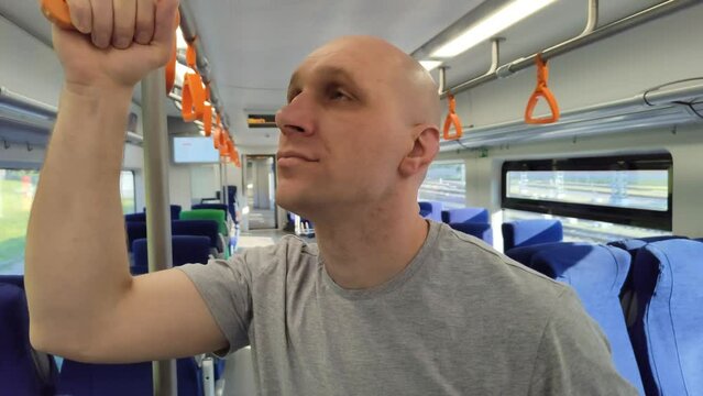 Young man rides train and looks around. Bald guy holding on to handrails in subway or train car. Moscow Central Ring (MCC), Lastochka train. Passenger in empty carriage. Modern transportation system