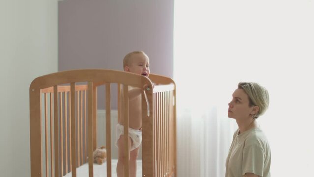 mother trying calm screaming crying baby toddler infant in crib put to bed cot or cradle. caucasian mother talking, petting, hugging hysterical child kid, persuading him to sleep. parenthood concept