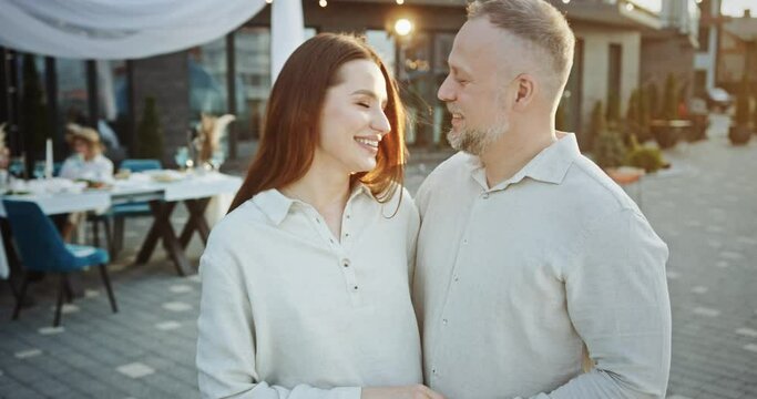 Loving positive couple hugging and kissing in outdoor restaurant. Enamored adult bearded man with happy wife in elegant outfits cuddling kissing and talking to each other while standing near table in