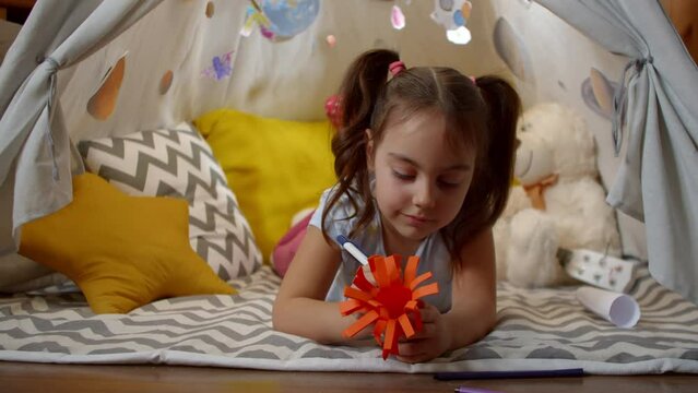 little girl makes crafts with her hands lying on the floor in a tent at home. Child is concentrated on decorating a paper toy. Concept of education and training of children.