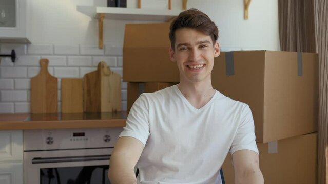 POV of happy young man looking in camera sitting at glass table near closed laptop in kitchen. Guy touches neck with hand against paper boxes slow motion