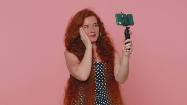 Redhead woman in dress traveler blogger taking selfie on mobile phone selfie stick, communicating video call online with subscribers. Young ginger girl isolated alone on pink studio background