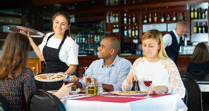 Attractive young hispanic waitress serving delicious pizza to friendly company in restaurant